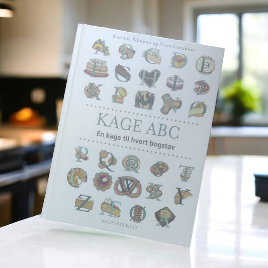 Hvid bog i et køkken og forsiden af bogen er fyldt med alfabetet og billeder af kager som passer til alfabetet fx L og en lagkage